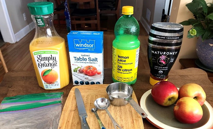 Ingredients are displayed on a tabletop, along with a cutting board and utensils. In the back, there is a jug of orange juice, a box of table salt, a bottle of lemon juice, and a squeeze bottle of honey, as well as three red and yellow apples on a plate to the side.