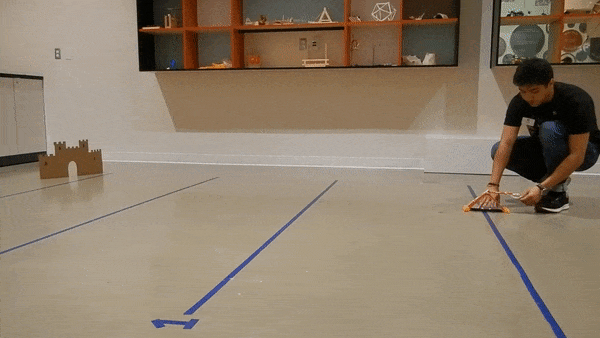 A cork is fired from a K’NEX catapult towards a cardboard target. The cork falls far short, rolling across the floor. 