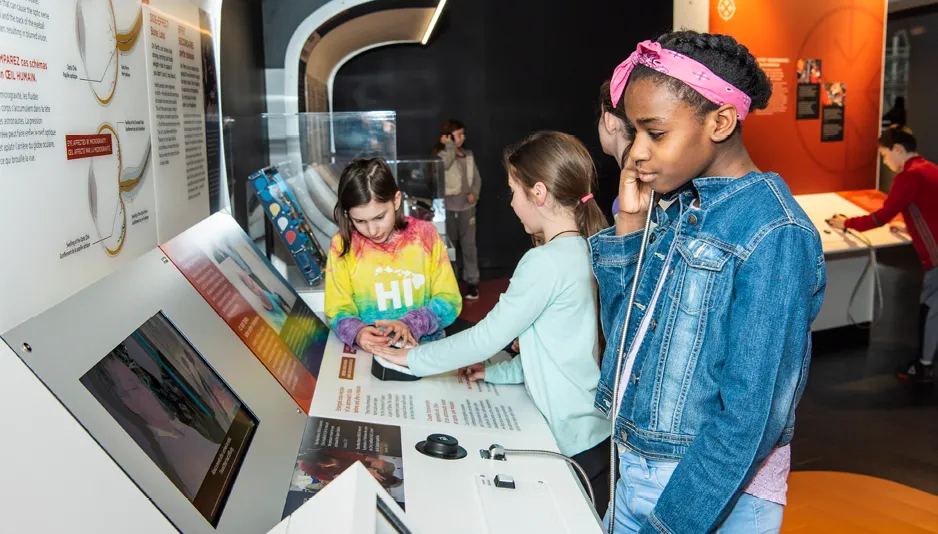 Three children look at a museum exhibit interactive panel.  The child in the foreground, wearing a jean jacket and pink headband listens on an earphone. The other two children in the background examine a visual panel