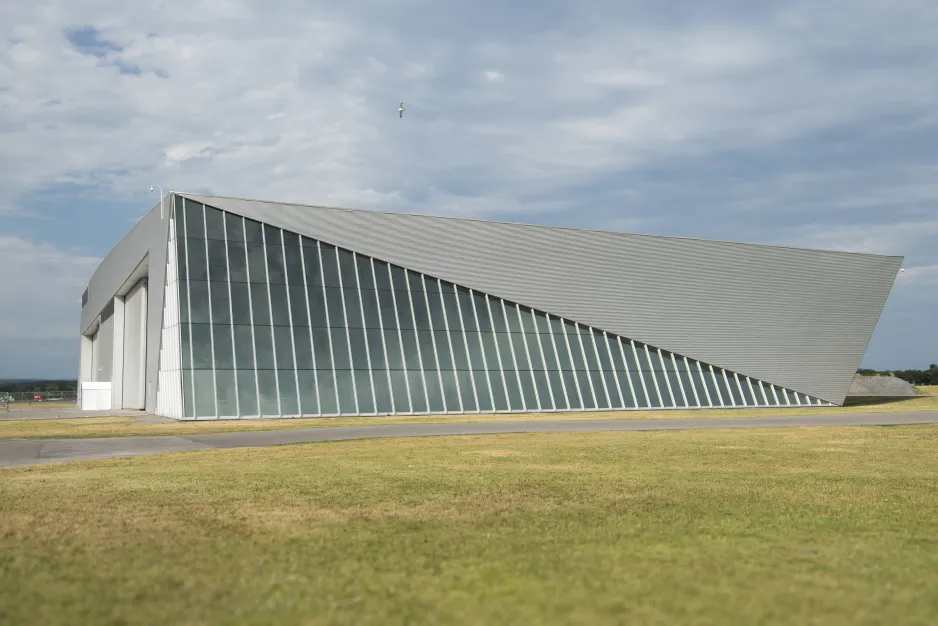 Photo of the museum's Reserve Hangar, a large, grey building with  a large triangle of windows along the front.  The building is set against a blue sky dotted with clouds and a field of green grass.