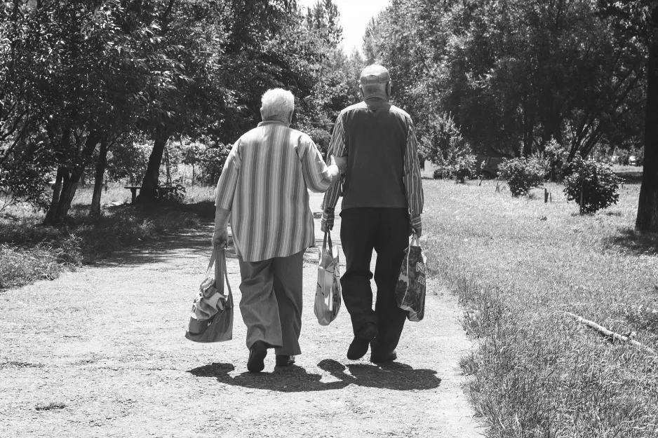 Un couple de personnes âgées se promène dans une ruelle arborée portant leurs courses