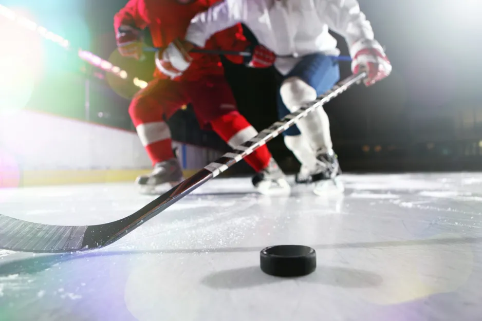 Image prise au niveau de la glace et montrant une mise en échec. On y voit les bas blancs et la culotte bleu foncé de l’un des joueurs, ainsi qu’un bâton avancé vers la rondelle.