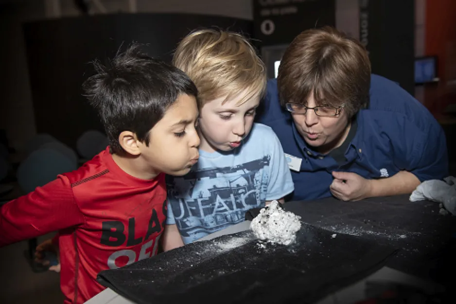 A female museum guide and two young boys blow on a ball of dry ice sitting in front of them.