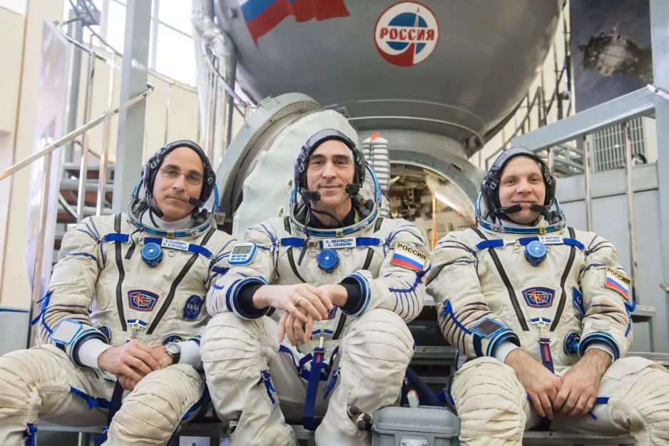 The three astronauts set to launch on April 9 in space suits, in front of their launch vehicle a Soyuz spacecraft