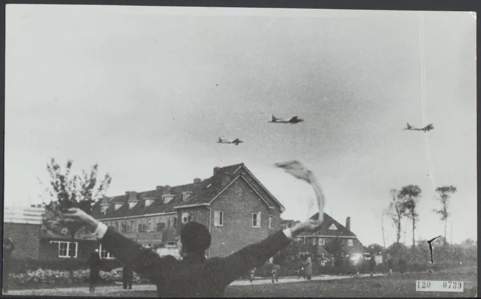 A black-and-white image shows a person waving a white cloth overhead while watching planes fly low over a residential area. Wikimedia Commons