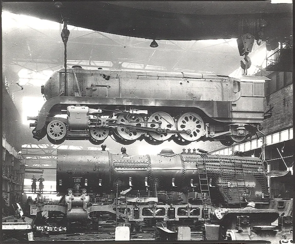 Photographie en noir et blanc d’une locomotive à vapeur en construction. Elle montre une locomotive suspendue dans les airs avec des pièces au sol.