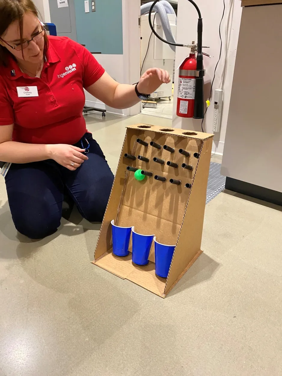 A woman in a bright red Ingenium shirt sits on the floor next to a cardboard ball drop game. Her aim is to put a green ball through one of three holes at the top of the cardboard structure so the ball can travel through a series of mini poles to land in one of three blue cups at the bottom.