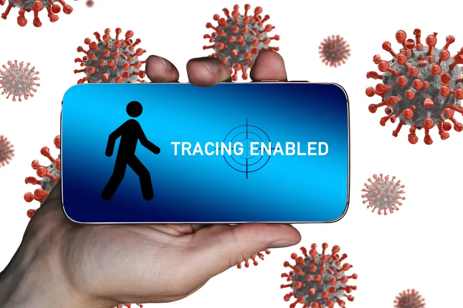 A hand holds a smartphone with the words ‘Tracing Enabled’ on the screen. Multiple animated representations of the COVID-19 virus are in the background.
