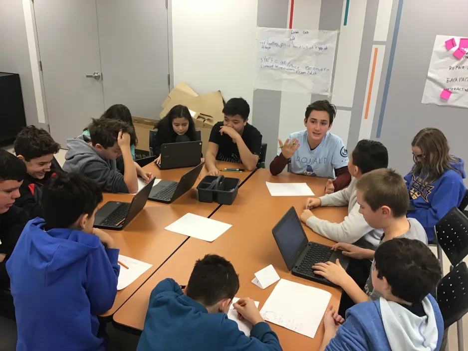 A group of 13 students gather around a table. They are discussing ideas and planning the projects. Some speak, some draw, and some do research on laptops