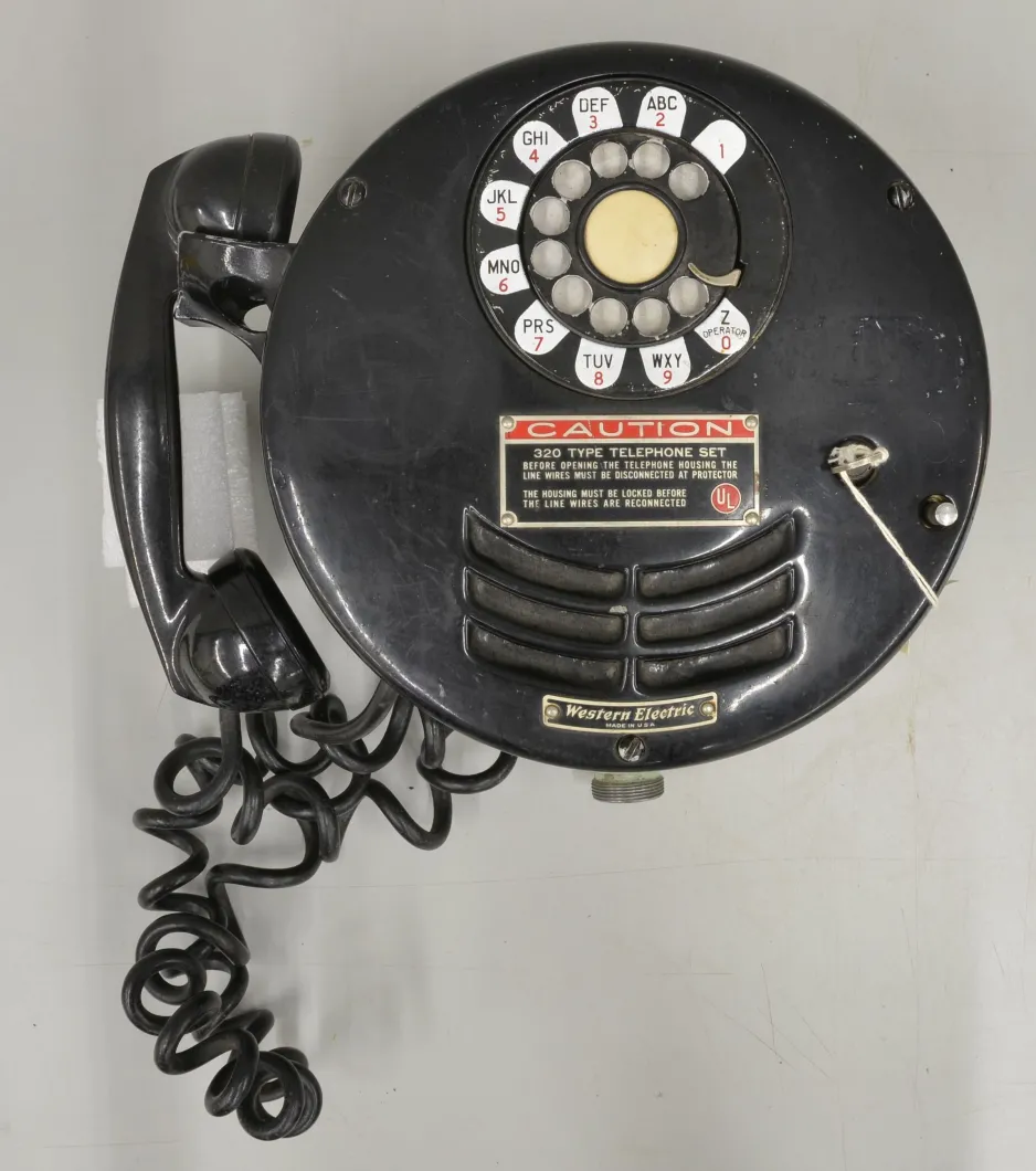 A round, black coated, metal rotary mine phone sits on a grey background.