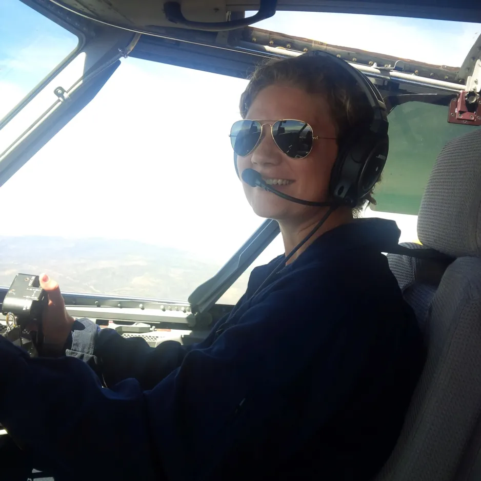 A young woman wearing a uniform, sunglasses, and a headset sits in the cockpit of an aircraft.