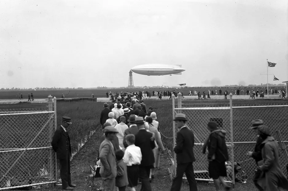 A black and white photograph of a crowd of people streaming through a gate, with a massive blimp-like airship tied to the top of a tall tower that looks like a lighthouse in the background.