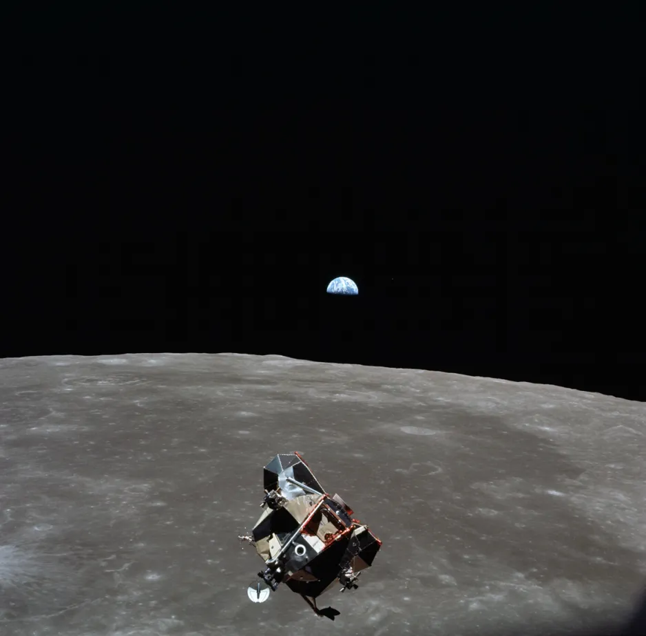 Vue de l’étage de remontée d’Apollo 11 en avant-plan, survolant la Lune grise. Au-dessus de l’horizon de la Lune, on voit partiellement la Terre dans l’ombre de la Lune contre l’espace noir (le lever de la Terre).