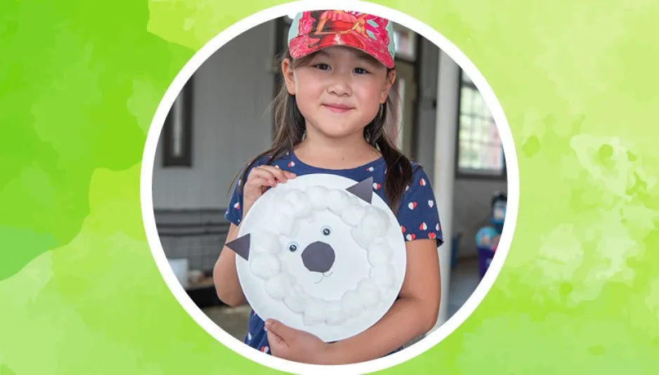 A girl smiles as she holds up a paper plate sheep craft, she is inside a circle against a lime green background.