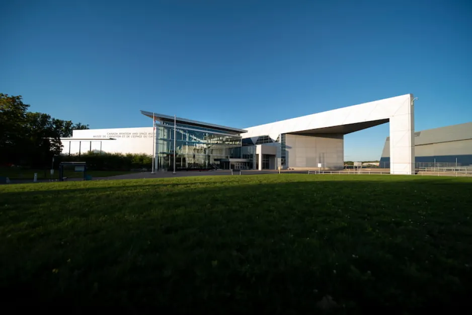 La façade angulaire d’un grand bâtiment blanc, avec les mots « Musée de l'aviation et de l'espace du Canada » visibles sur son côté gauche. De l’herbe verte est visible en avant-plan et un ciel bleu et dégagé en arrière-plan.