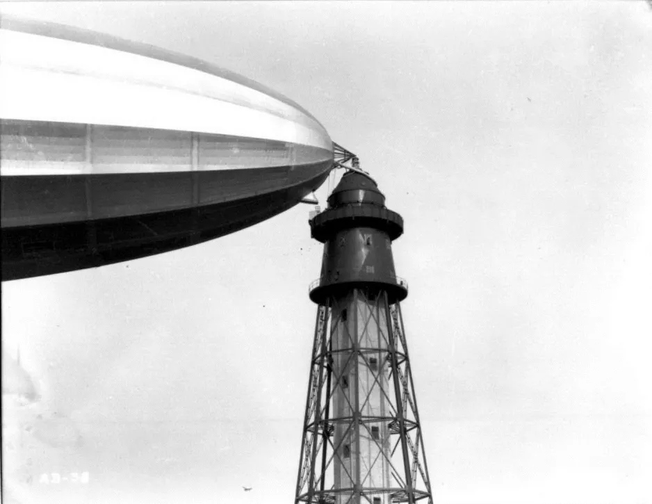 A black-and-white photograph showing a close-up view of the tip of a massive blimp-like airship, which is tied to the top of a tall tower that looks like a lighthouse.