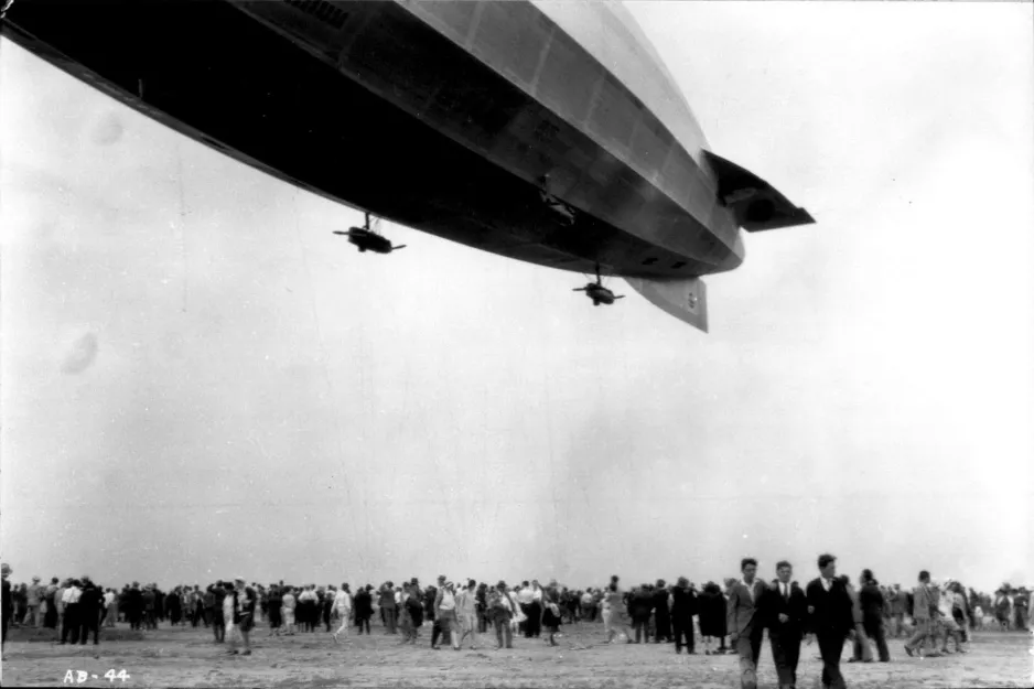 A black-and-white photograph showing a crowd gathered on the ground, below the back half of a massive blimp-like airship. Two small engine nacelles are attached to the underside of the airship.