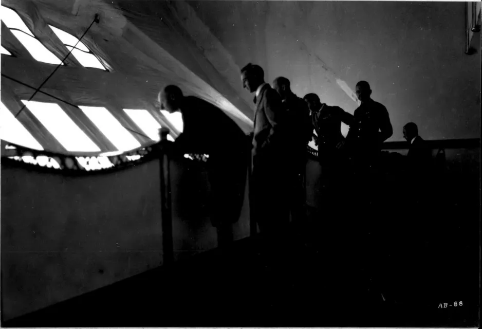 A black-and-white photograph of six silhouetted figures inside of the R-100 airship, looking out of a series of small, square windows.