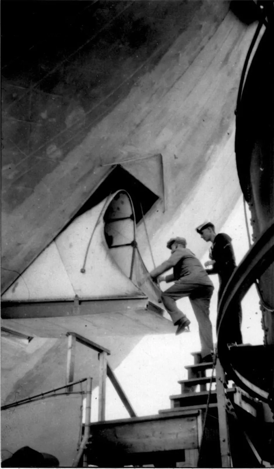 A black and white photograph of a man climbing onto the entrance of the R-100 airship from a set of stairs. The gap is approximately 1 metre tall. Another man stands behind him.