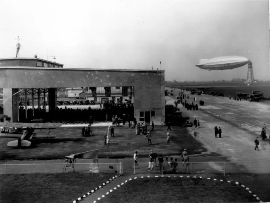 A black and white photograph of an airport building and runway, showing the massive blimp-like airship tied to a tall tower-like structure in the background.