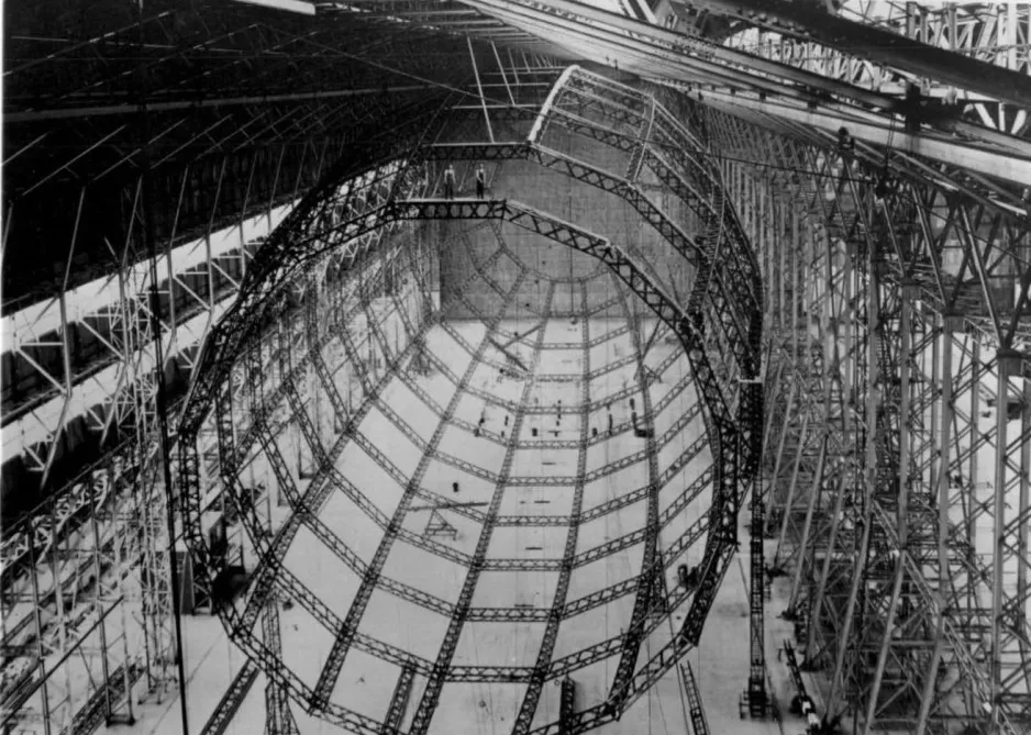 A black-and-white photograph of a massive bird cage-like structure made of aluminium beams, suspended on scaffolding.