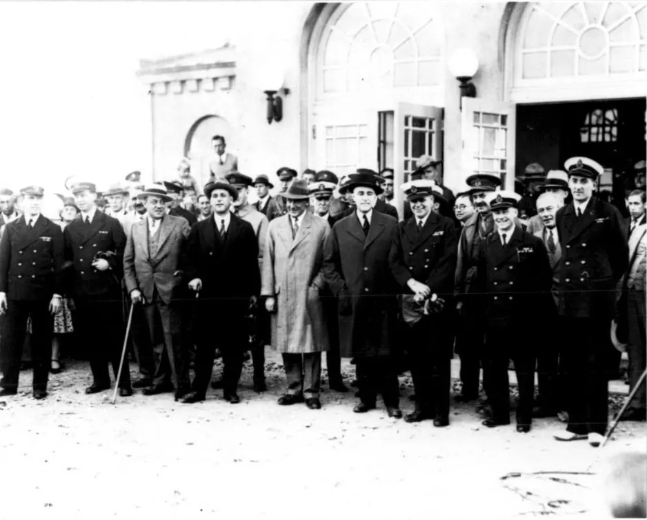 A black and white photograph of a group of men, some wearing military uniforms, others wearing suits and long coats, posing at the arrival event.