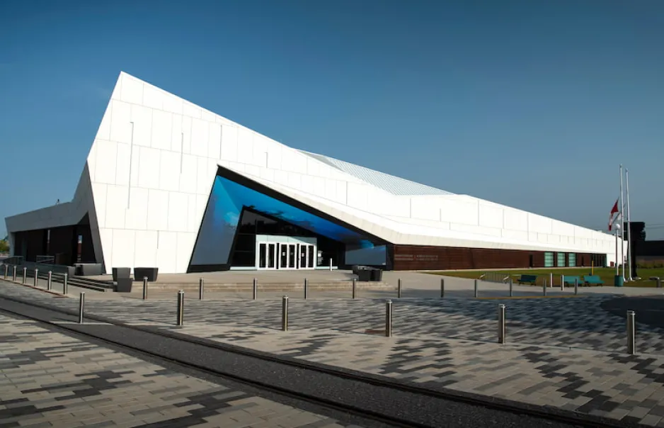 La façade d’un large bâtiment blanc géométrique se détache sur un ciel bleu et dégagé. Autour et au-dessus des portes d’entrée du bâtiment, un grand écran affiche des tons bleus éclatants.