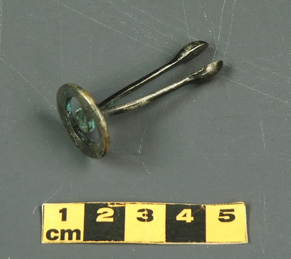 Petit dispositif contraceptif en métal, avec une base circulaire plate et ajourée, muni de deux bras parallèles, joints à la base, et de têtes ovales coupées en deux. Cet artefact mesure 5 cm de long.