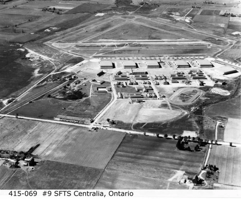 A black-and-white image depicts an aerial view of an RCAF base. It shows the buildings as well as the airstrip of the base surrounded by farmland.