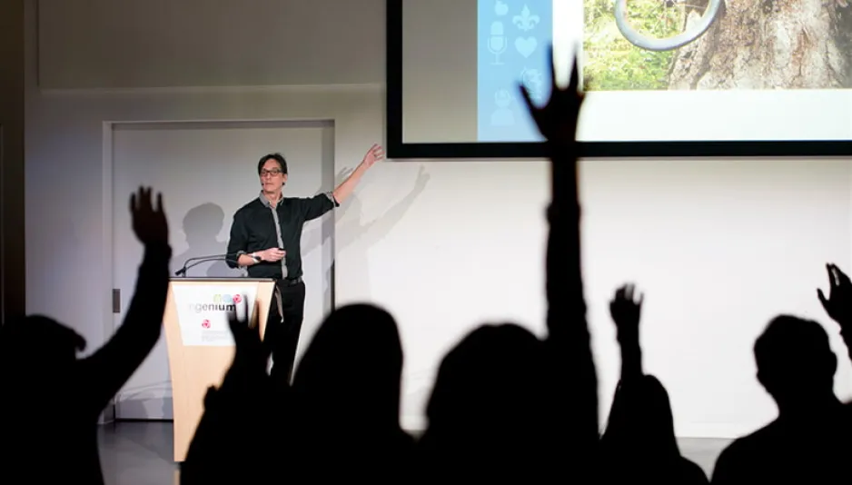 Photo of a speaker on stage pointing to a powerpoint presentation