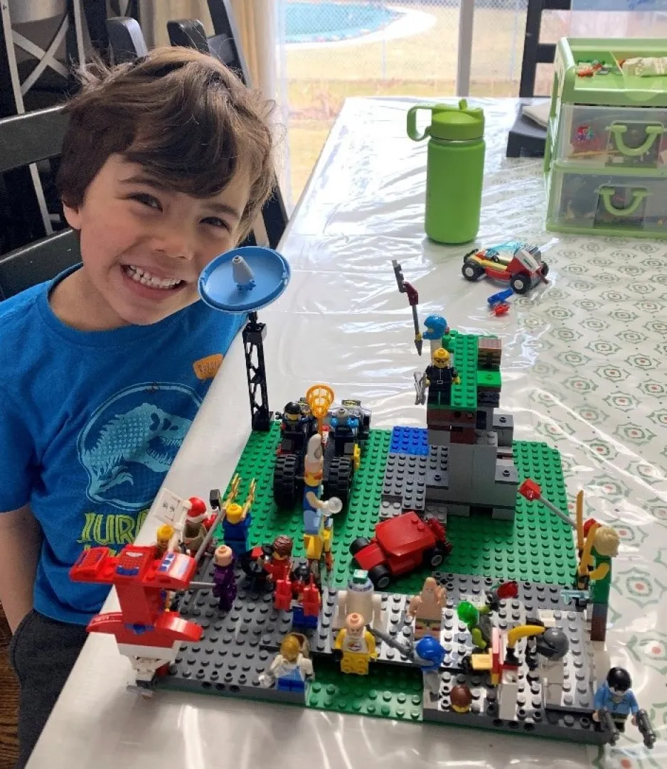 A little boy wearing a blue t-shirt smiles widely as he sits next to a table. On the tabletop is an elaborate and colourful LEGO creation.