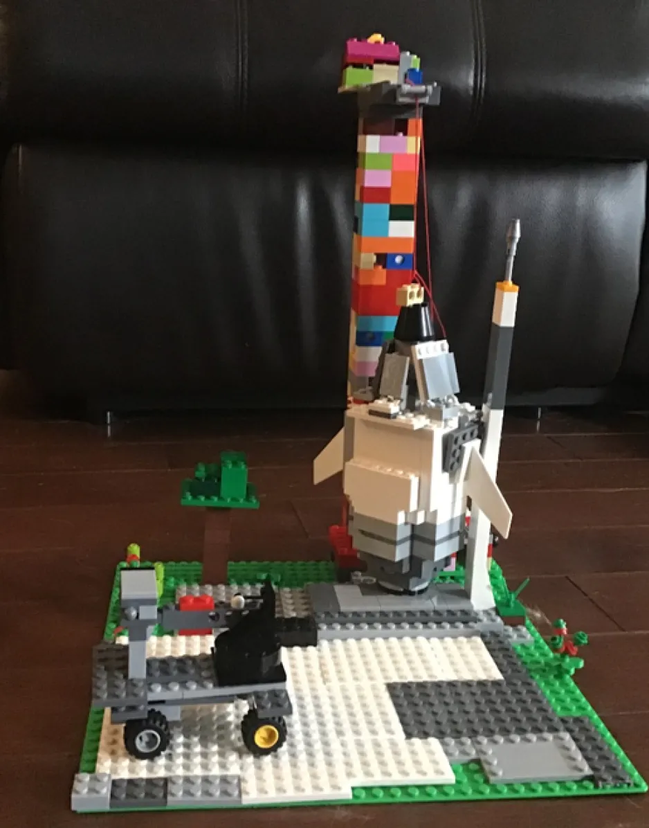 A LEGO creation that includes a rocket ship, a tower and launchpad, and a moon rover. The creation is standing upright on a wooden floor with a black background.