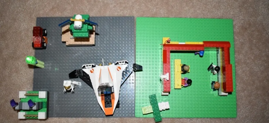 An aerial view shows a LEGO creation on the floor. A rocket ship made of LEGO is visible, and several LEGO people sit inside a red 