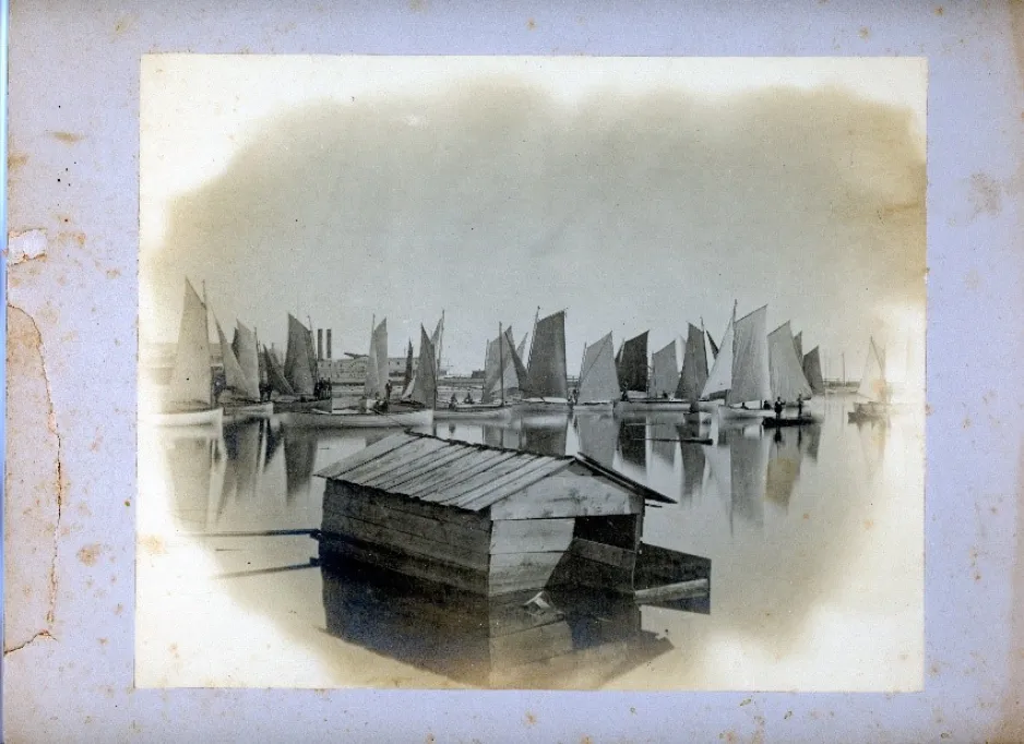 A fleet of sail-powered fishing boats clustered in harbor with a well-worn wooden boathouse in the foreground.