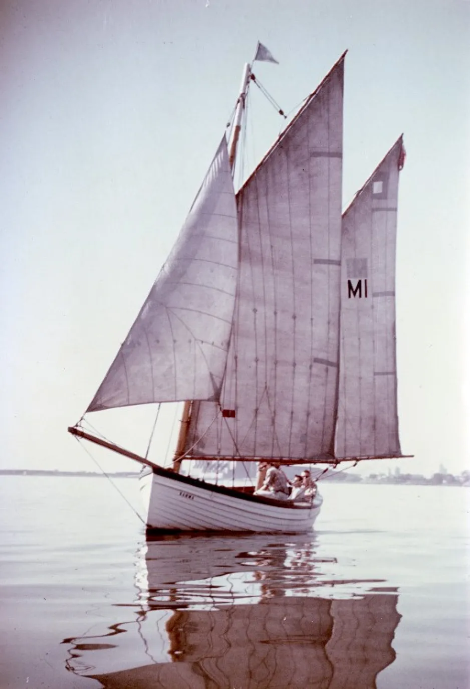 A white wooden boat with a large sails floats on the calm surface of a lake; three people are visible inside the boat.