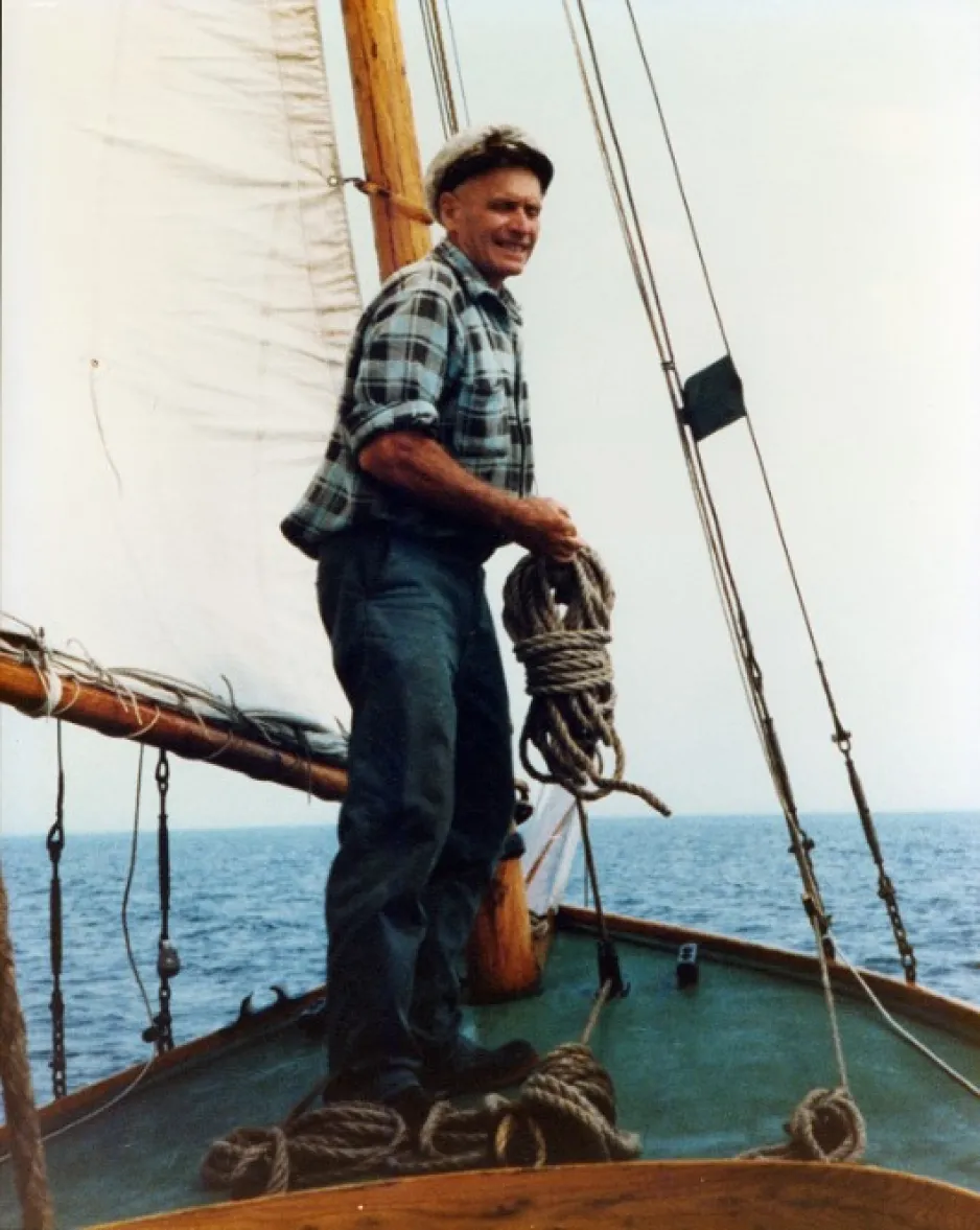 A man wearing a sailor’s hat stands on the deck of a boat holding a rope; the boat is surrounded by open water. 