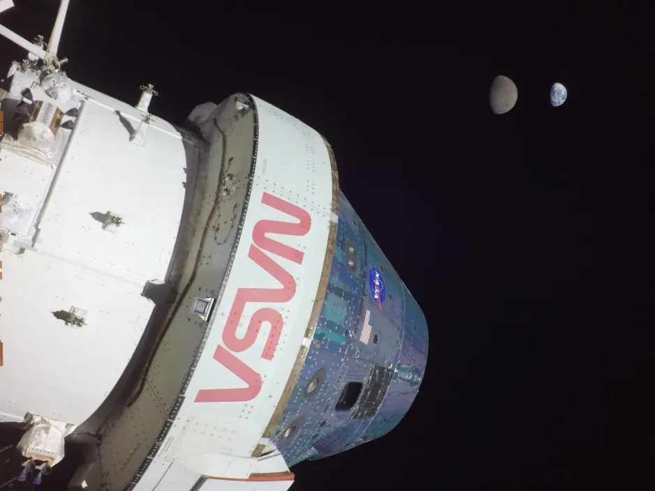 The Orion spacecraft in the left foreground, with a view in the background of the Earth and Moon, both partly in shadow against the black of space.
