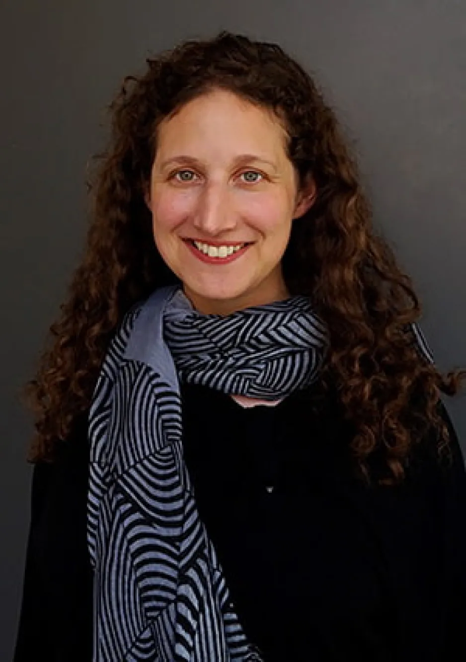 Dr. Rebecca Clare Dolgoy. A young woman with dark, curly hair smiles widely at the camera. She is wearing a dark shirt and a striped blue scarf.