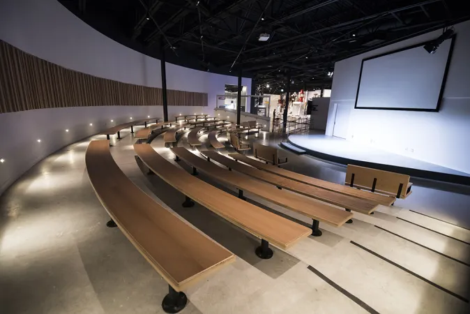Curiosity on Stage at the Canada Science and Technology Museum. The room features a semi-circle stage with a projector screen. Rows of wooden benches are curved around the stage with multiple stairs leading up to the last row of benches. There are a few seating options at the front before the start of the stairs.