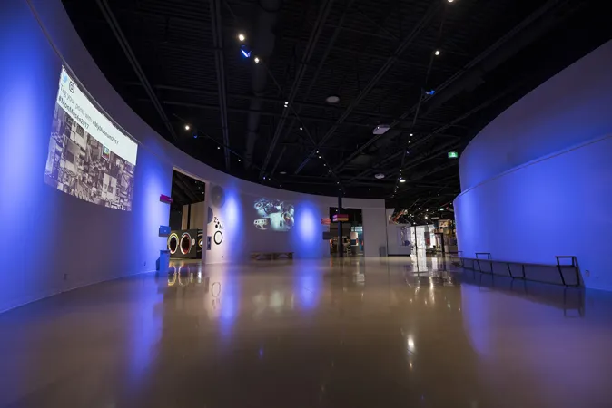 A large empty room in the middle of the Canada Science and Technology Museum. The walls are curved and white, the ceiling is high and painted black with cool-toned blue and white spotlights shining down. A slide is projected onto the side of one wall.
