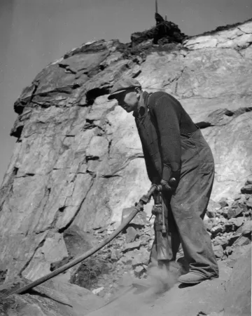 Breaking up large blocks of iron ore blasted from the face of a hill in Eastern Canada with an air drill, so it will pass through the crusher.