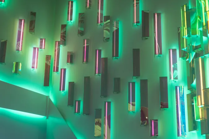A close-up of the lights at the entrance of the Canada Science and Technology Museum. The wall has an array of neon or fluorescent lights and mirrors in differently sized rectangles arranged vertically giving a kaleidoscope of reflections and colours. Currently, there are some magenta lines with glowing green, yellow, and aquamarine background lights.