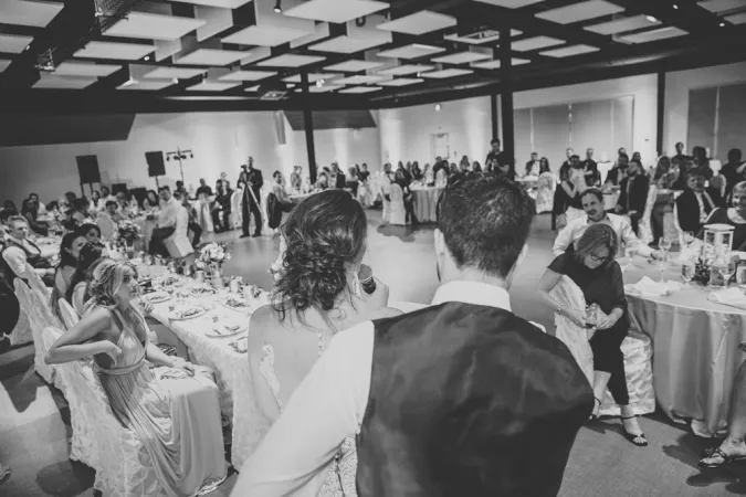 A black and white photo of what looks like a wedding. The back of two people’s heads can be seen making a speech while the audience listens attentively. The ceiling is dark but with large light-coloured squares hanging down giving the room a large spacious and airy feeling. 