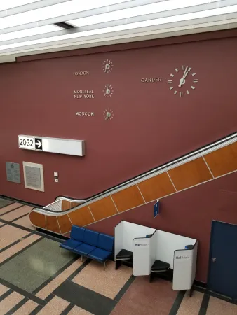 The escalator in the international terminal area with four clocks on the wall behind it giving the time of day in Gander, London, New York, and Moscow.