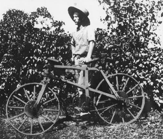 Photo noir et blanc montrant un homme debout, à l’extérieur, la main gauche posée sur le siège d’une bicyclette de bois.
