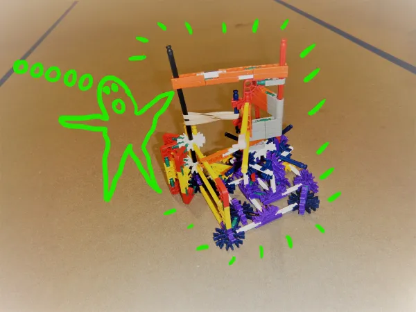 A completed sideways K’NEX catapult that launches a projectile from the side, instead of over the top. The image has a cartoon drawing of a green monster standing next to the catapult. 