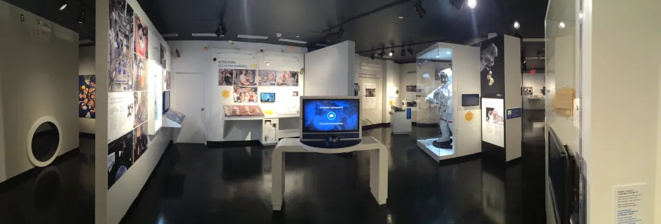 Vue grand-angle d’une grande salle de musée aux murs blancs et au plancher noir. On y voit des panneaux colorés et un écran d’ordinateur ainsi qu’une combinaison spatiale dans une vitrine en verre.
