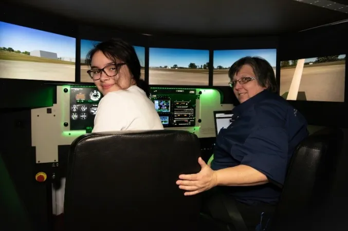 Two women smile as they turn to face the camera; they’re sitting inside a flight simulator.