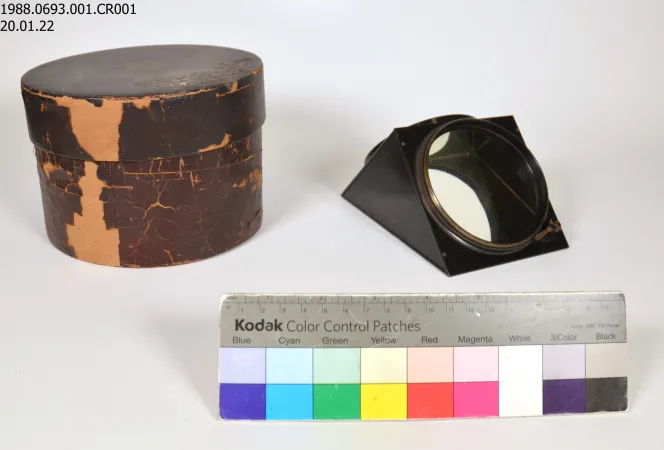 A small metal and glass prism and its old brown storage box sit on a white background. A Kodak color strip sits in front of the artifacts.  