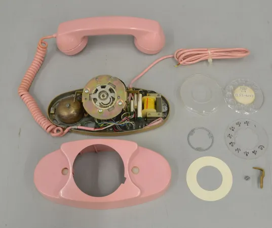A pink rotary phone with the plastic case removed and dial disassembled, sits on a grey background. 